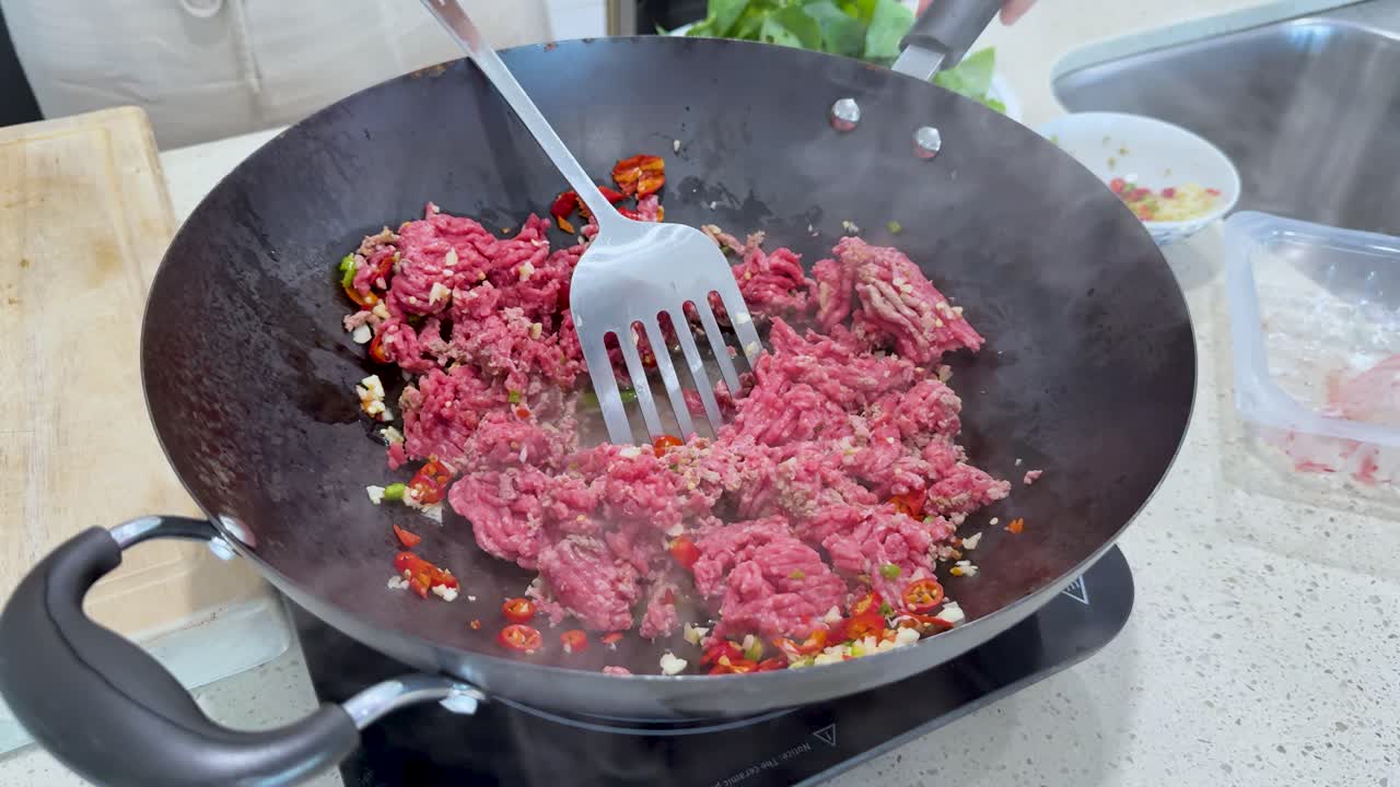 Raw minced meat is stir-fried with chopped chili peppers and vegetables in a wok, under bright kitchen lighting, with visible steam and utensil movement
