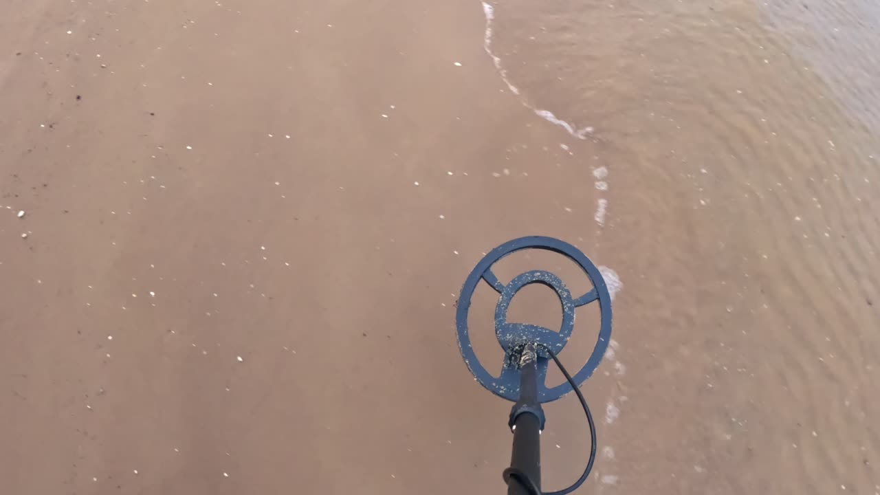POV sweeping a metal detector along the shoreline as gentle waves reach the sand, showing the focused search for lost objects near shallow water
