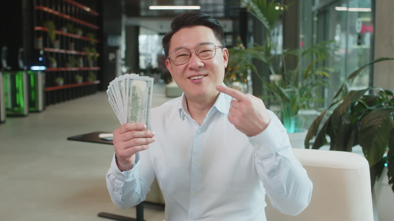 นักธุรกิจสูงวัยพร้อมธนบัตรดอลลาร์ ชูนิ้วโป้งขึ้นและมองมาที่กล้องในล็อบบี้สำนักงาน