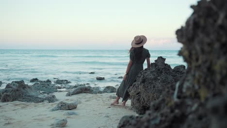 Frau-Steht-Am-Strand-In-Der-Nähe-Von-Felsen-Bei-Sanfter-Meeresbrise-Und-Trägt-Ein-Kleid-Und-Einen-Sonnenhut