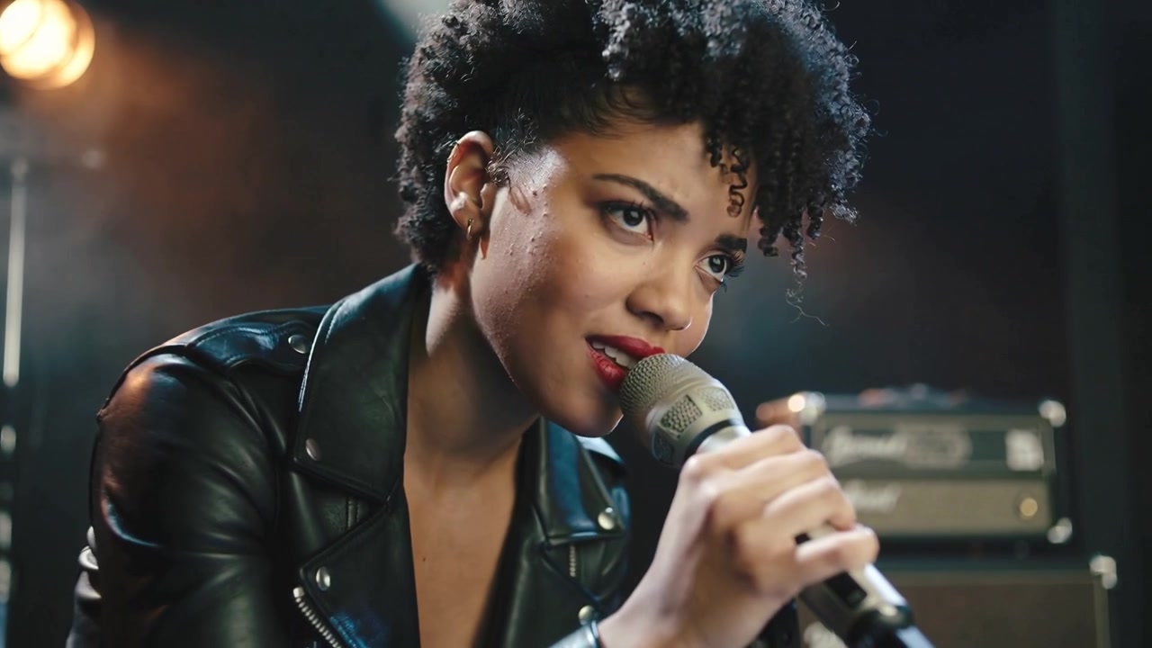 Performing singer leaning into mic after spotlight cue in studio, by amps wearing leather jacket