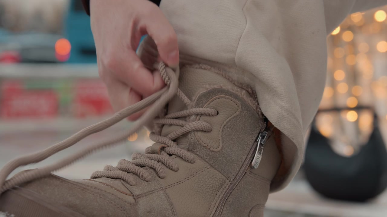 Un primo piano ad angolo basso mostra mani che tirano un laccio intrecciato e sistemano un nodo allentato su una scarpa da trekking in camoscio o tessuto color cuoio, mentre il risvolto di un pantalone beige poggia sopra il marciapiede, catturando una breve pausa e un momento all'aperto quotidiano.
