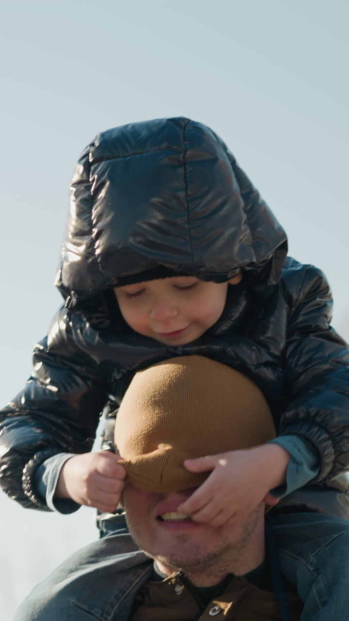 un primer plano de un padre llevando a su hijo pequeño en sus hombros mientras camina al aire libre, el hijo ajusta juguetón la gorra del padre, que cubre el rostro del padre