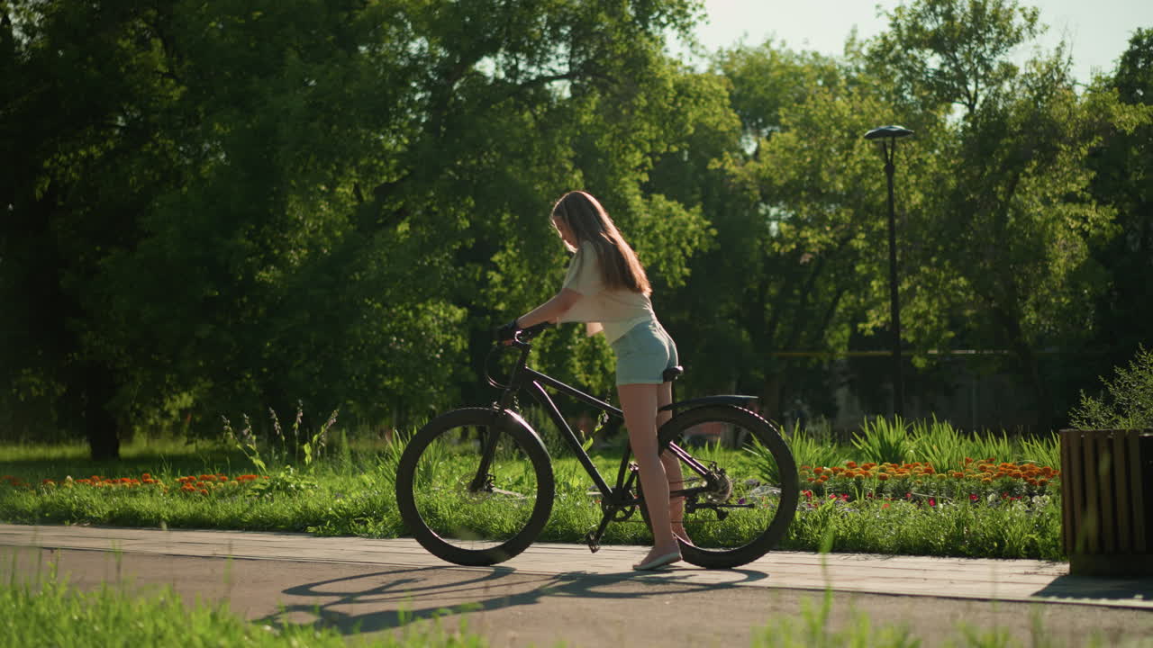 여성 자전거 타는 여성은 다채로운 정원 근처에 자전거를 타고, 배경에는 울창한 초록색, 나무, 그리고 따뜻한 빛 아래 활기찬 꽃병이 있습니다.