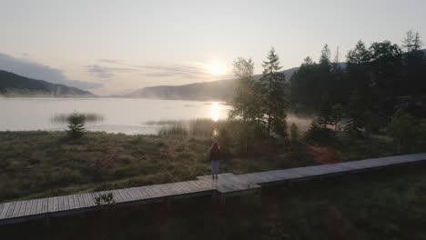 drone overtake shot of a young woman watching a sunrise on a misty lake