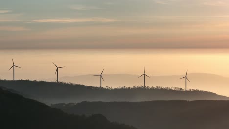 rotating wind turbines on forest-covered hills at sunset in portugal. drone footage, moving slowly upwards.