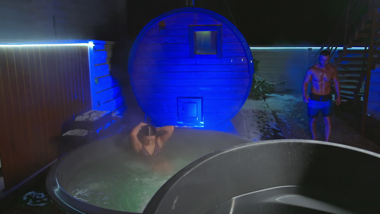 Good-looking Caucasian woman in stripy swimsuit sits in a hot tub outdoors in winter. Man wearing shorts appears, comes up and deeps into the barrel with cold water gasping.