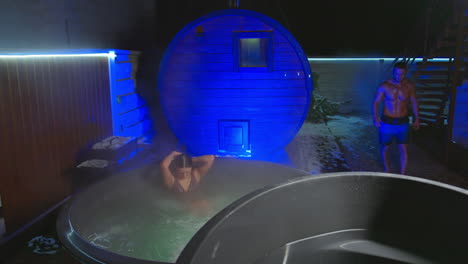 good-looking caucasian woman in stripy swimsuit sits in a hot tub outdoors in winter. man wearing shorts appears, comes up and deeps into the barrel with cold water gasping.