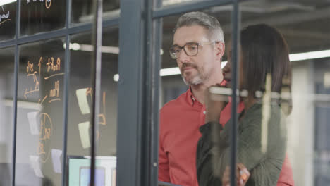 focused diverse male and female colleague brainstorming writing on glass wall in office, slow motion