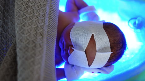 little infant lies in bed holding hands behind head. baby has a bandage on eyes for protection from uv light. jaundice treatment. top view.