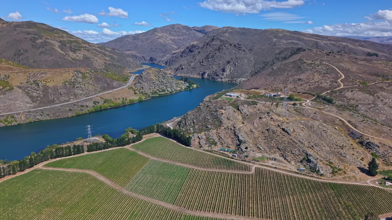 A drone ascends backward over neat vineyard rows near Cromwell, passing above power lines and houses, revealing the Kawarau River and distant Clutha Valley mountains