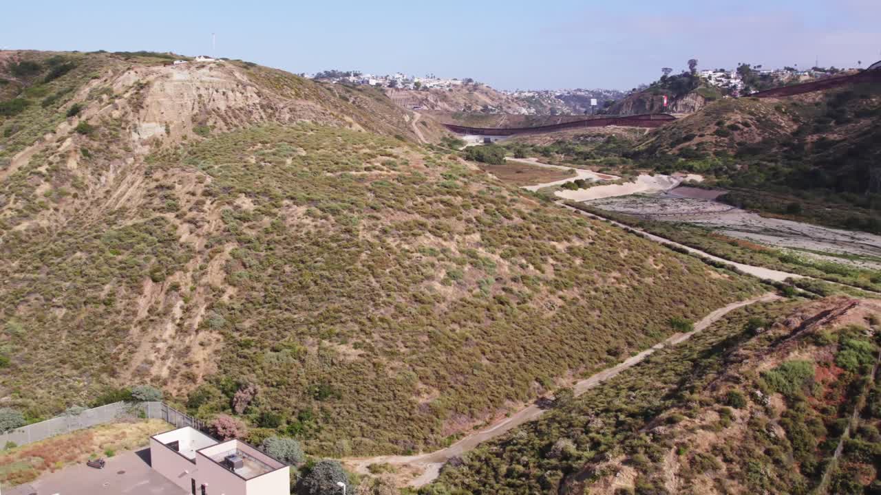 Aerial footage showcases dry, rugged hills with a winding road and distant views of the U.S.-Mexico border wall and residential developments under clear skies.