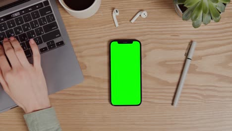 person typing on laptop with green screen phone on wooden desk