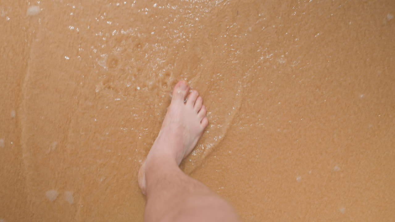 Bare feet stepping into foamy surf on golden sand, closeup view of toes, footprints and gentle wash intimate shoreline moment showing wading motion, soft bubbles, tactile sand texture and calming