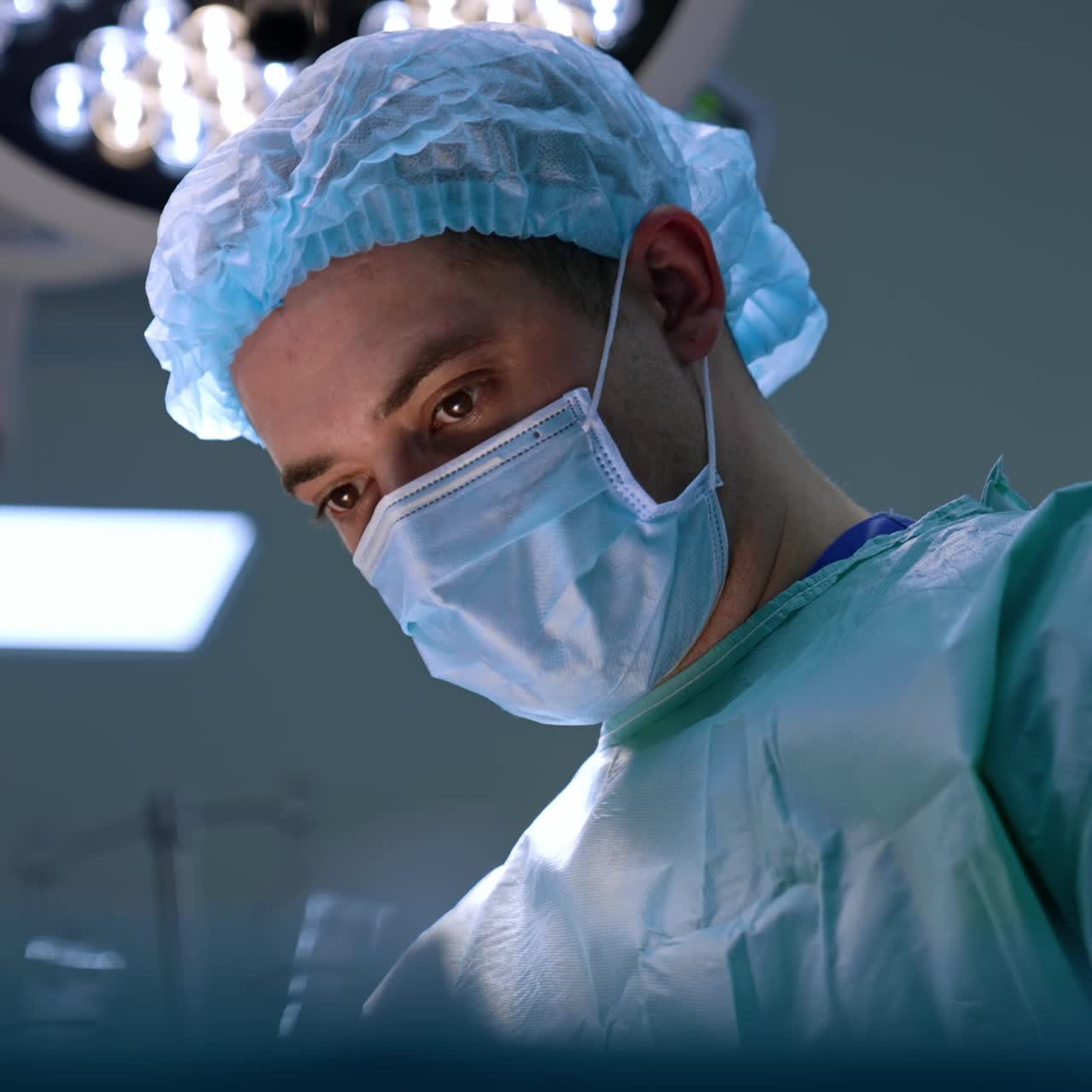 Primo piano del viso concentrato di un chirurgo caucasico di mezza età che indossa mascherina e cuffia. Inquadratura dal basso. Ritratto di un medico che esegue una procedura in una sala operatoria moderna.