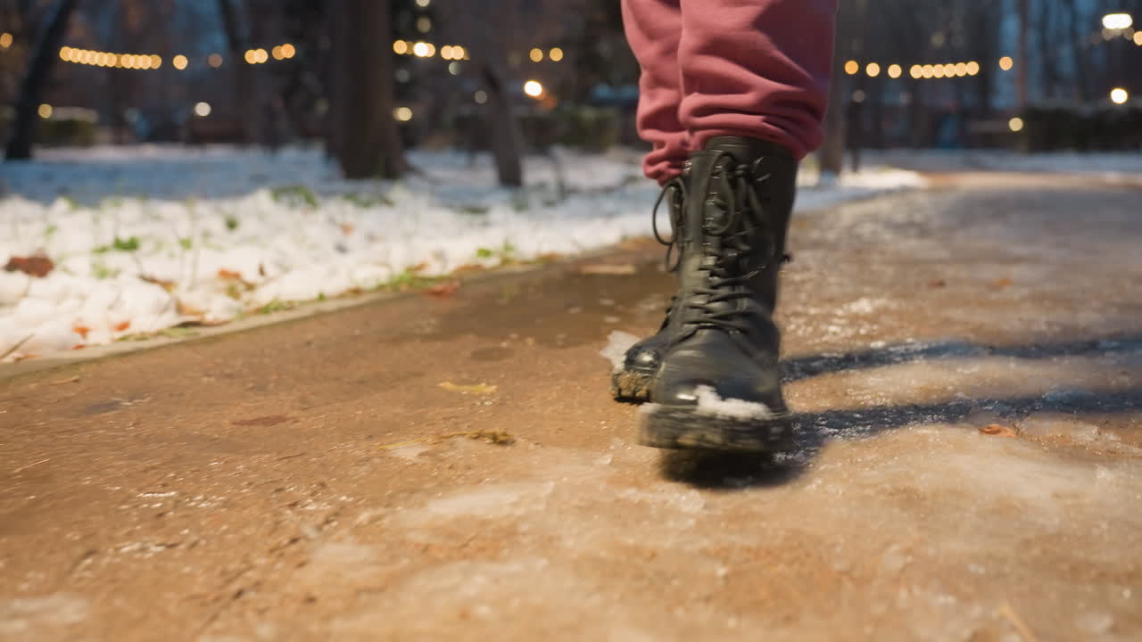 primer plano de la bota pisando un sendero cubierto de nieve en el parque de invierno. suelo nevado, clima frío, luces de la calle iluminadas creando una escena urbana de invierno en el parque de la ciudad