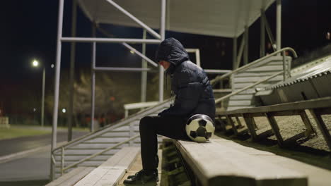 side view of a man in a black outfit and jacket sitting alone on a stadium bench with his hands between his legs, quietly, with a soccer ball beside him, under dim stadium lights