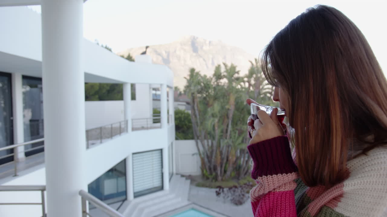 Drinking tea, young woman relaxing on balcony overlooking pool and mountains, copy space