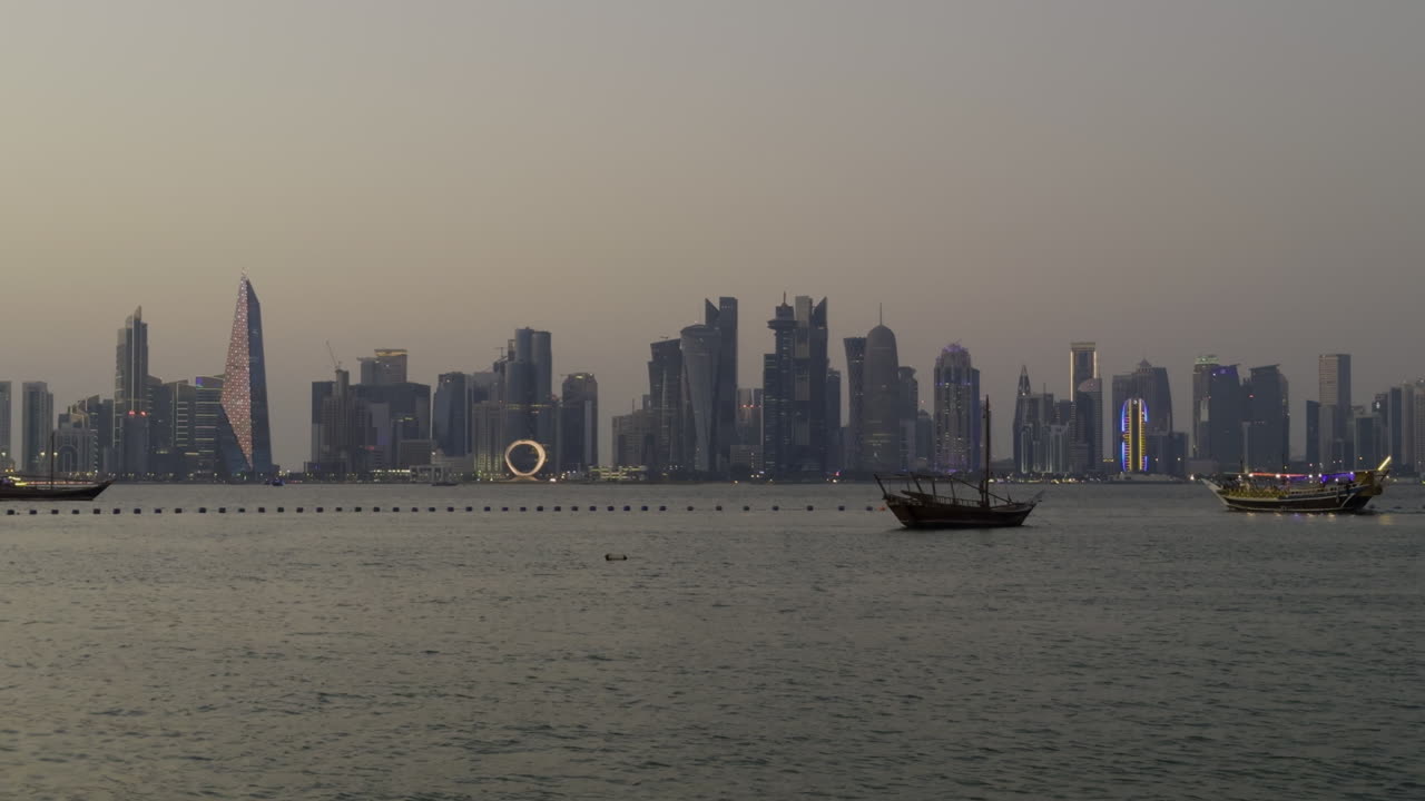 Doha skyline at sunset, featuring modern skyscrapers along the waterfront, with boats in the foreground as sunset glow spreads across sky on multi colored buildings