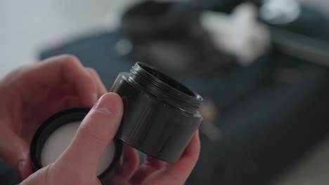 hands open cream jar scoop styling paste close jar then rub paste into palms in bright loft studio setting with suitcase and mirror in blurred background shot in natural light