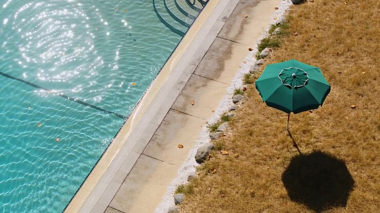 Sunlight shifting, green umbrella casting shadow on dry lawn by rippling turquoise pool, copy space