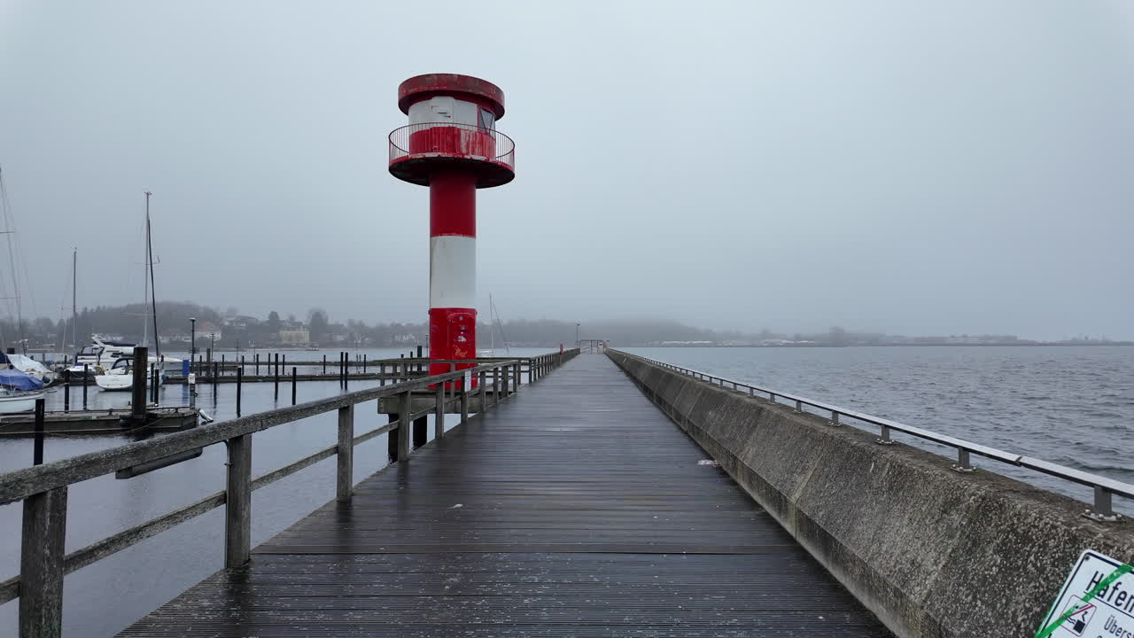 Vista panoramica del faro rosso e bianco all'ingresso del porto di Eckernförde, in Germania, immortalato in una giornata nebbiosa e nuvolosa. Un lungo molo di legno conduce verso il faro, con il Mar Baltico calmo