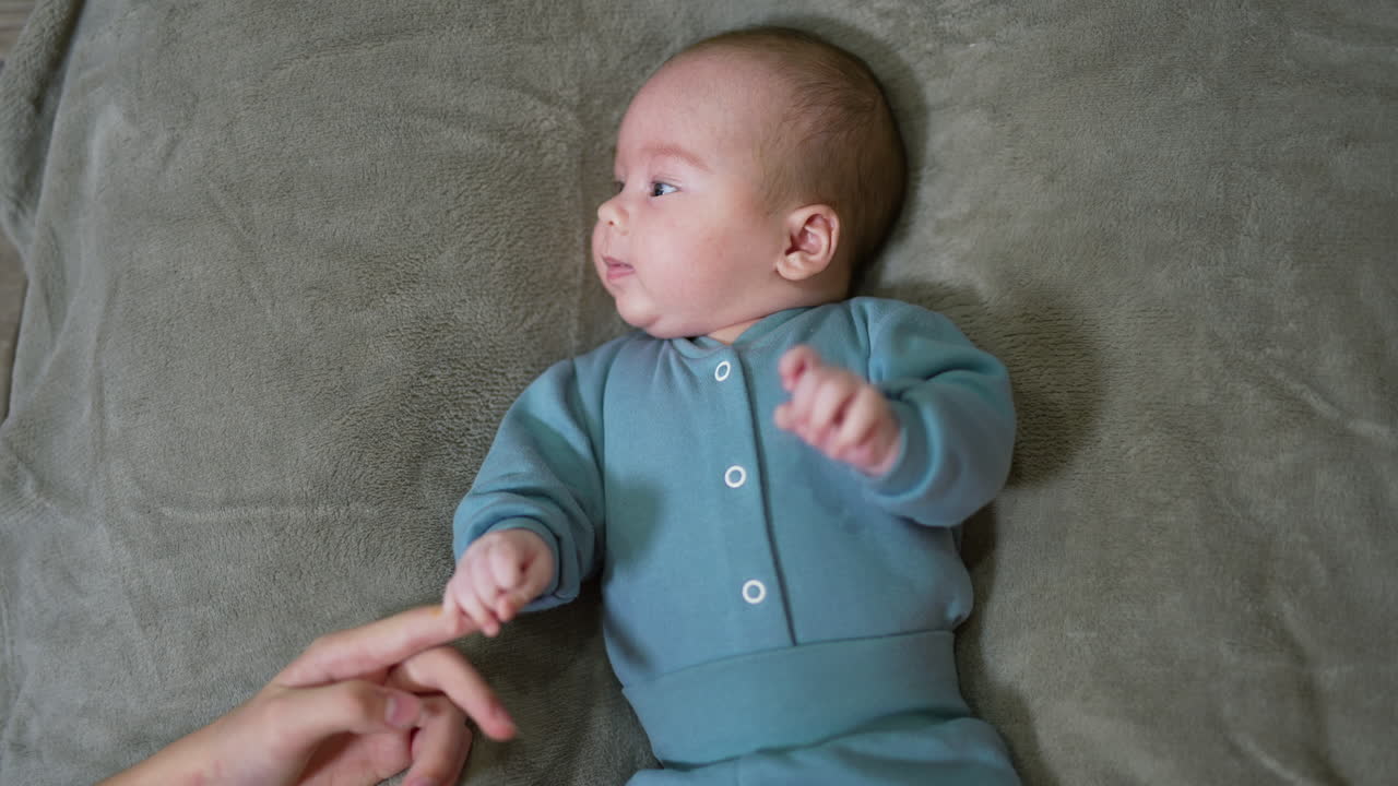 Lovely kid lying on the bed and tossing his arms and feet. Mommy stretches hand to child and he grabs her finger. Grey background.
