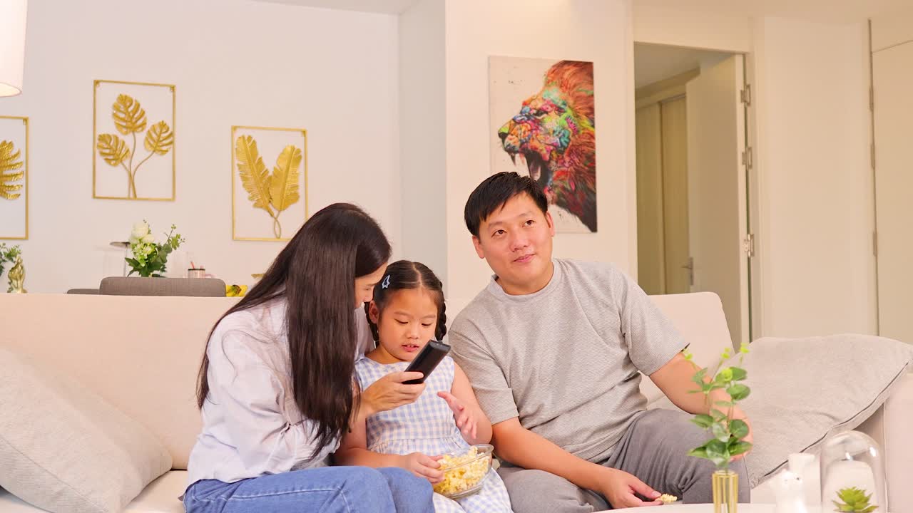 Parents and daughter smiling, eating popcorn, watching TV in bright, cozy living room setting