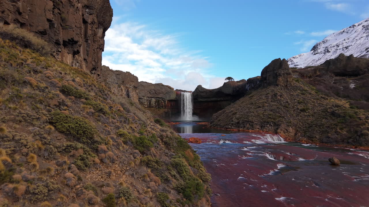 Salto del Agrio plunges into a basalt amphitheater framed by reddish volcanic rock terraces and snow-dusted peaks in Neuquén, Argentina, captured in dramatic aerial approach