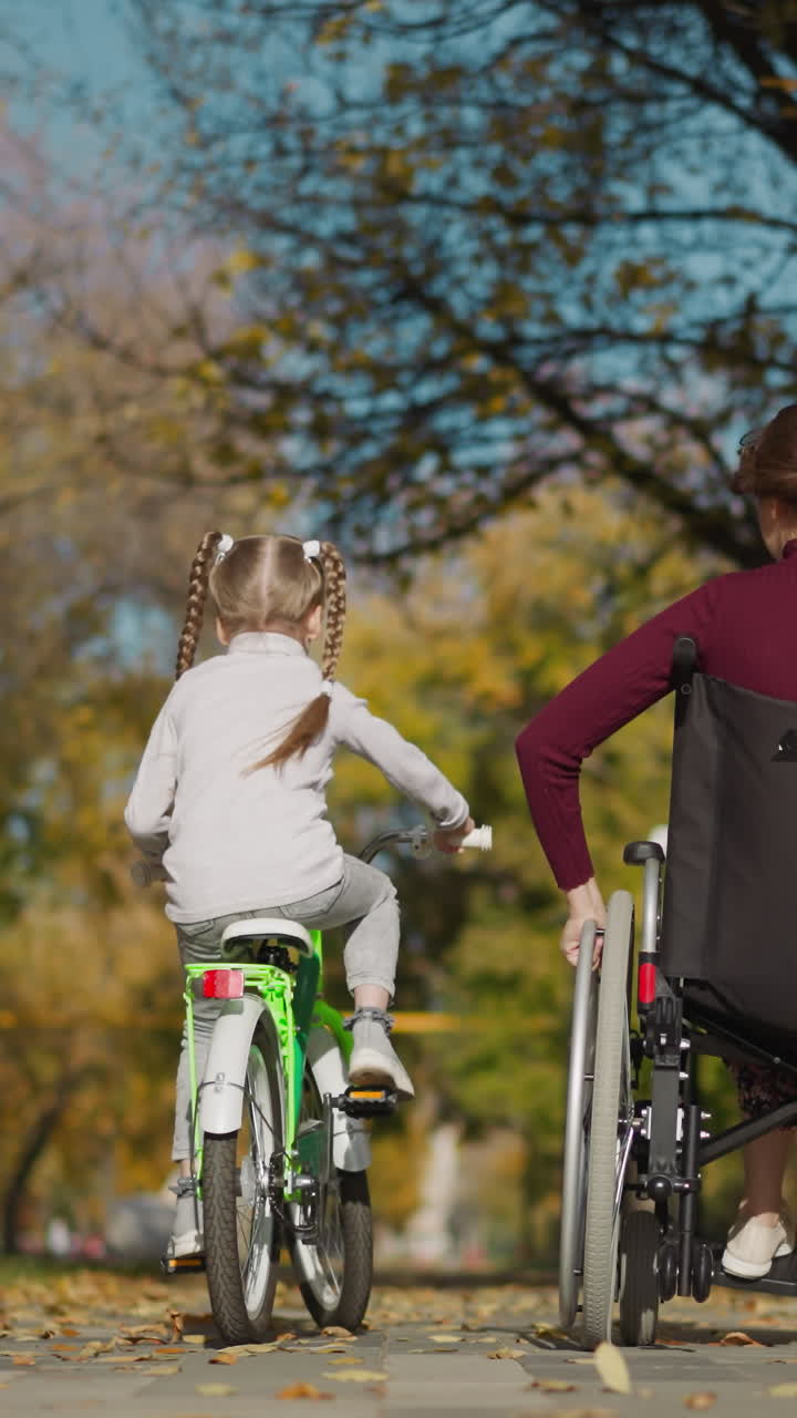 la hermana mayor sentada en silla de ruedas y la hermana menor en bicicleta se comunican moviéndose por el parque. vacaciones familiares en la naturaleza en un soleado día de otoño vista trasera
