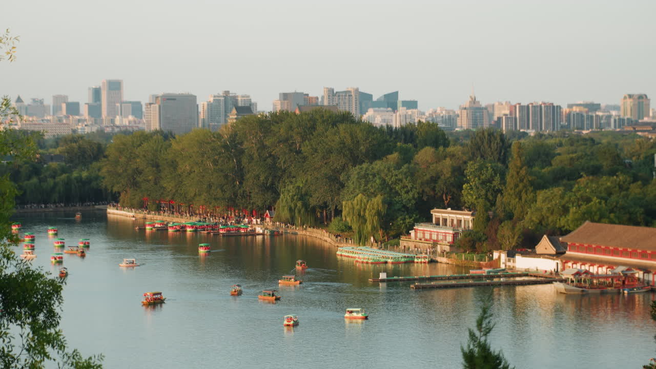 Miejskie jezioro z rowerkami wodnymi, zalesionym brzegiem, przystanią i panoramą miasta jaśniejącą w świetle późnego popołudnia, rodziny i turyści pływają wśród kolorowych jednostek, spokojna woda odbija miejski krajobraz, ciepłe lato.