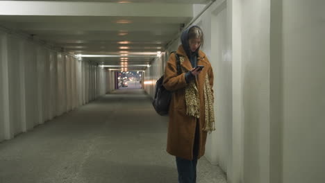 a girl in a brown coat and jeans walks through an underpass tunnel, leaning against the wall while looking at her phone with a worried and sad expression