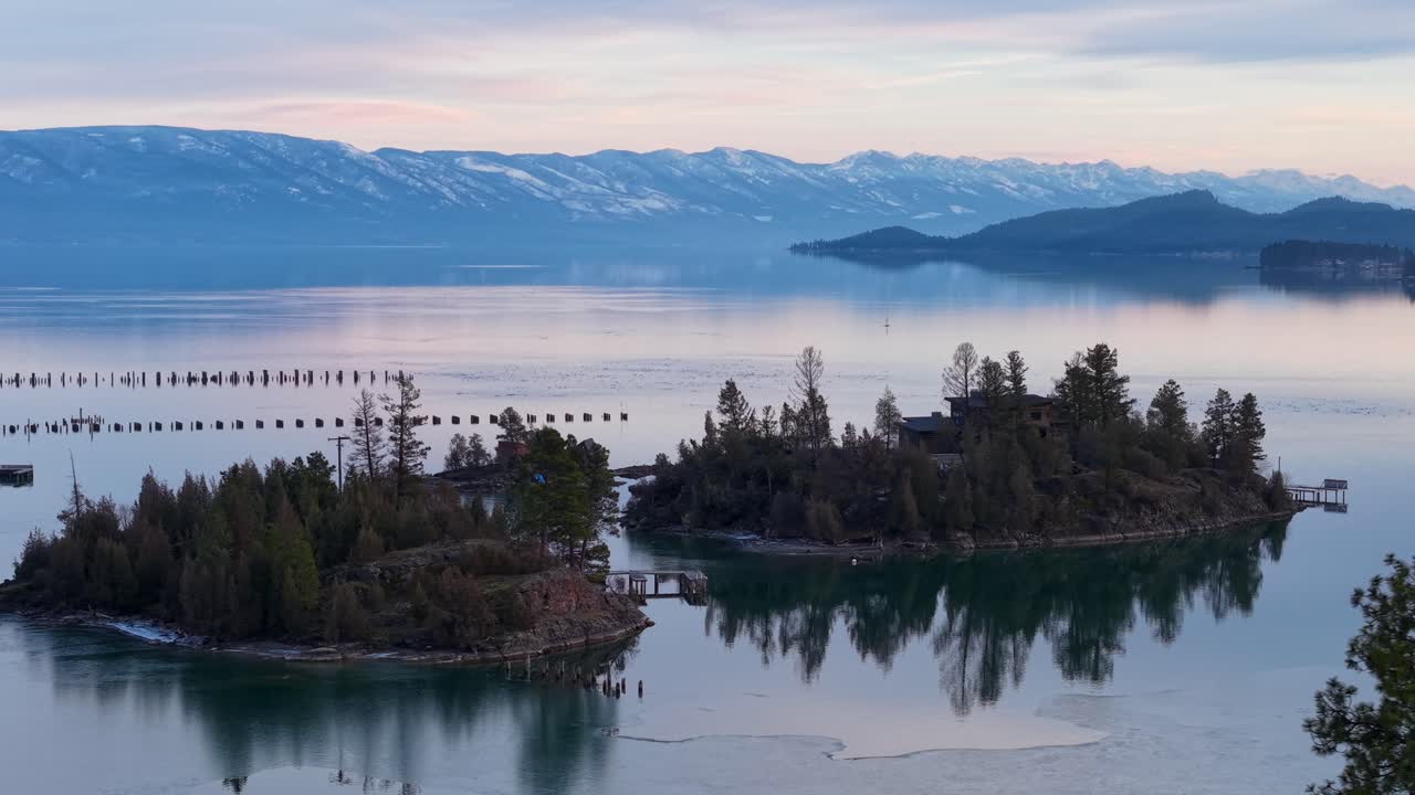 Riprese aeree di un lago tranquillo al tramonto con un gruppo di isole coperte da alberi e pali di legno lungo la riva, con montagne sinuose sullo sfondo sotto un cielo tenue e pastello