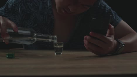 술에 취한 우울한 아시아 남자가 스마트폰을 사용하는 동안 유리잔에서 보드카를 마시고 있습니다.
