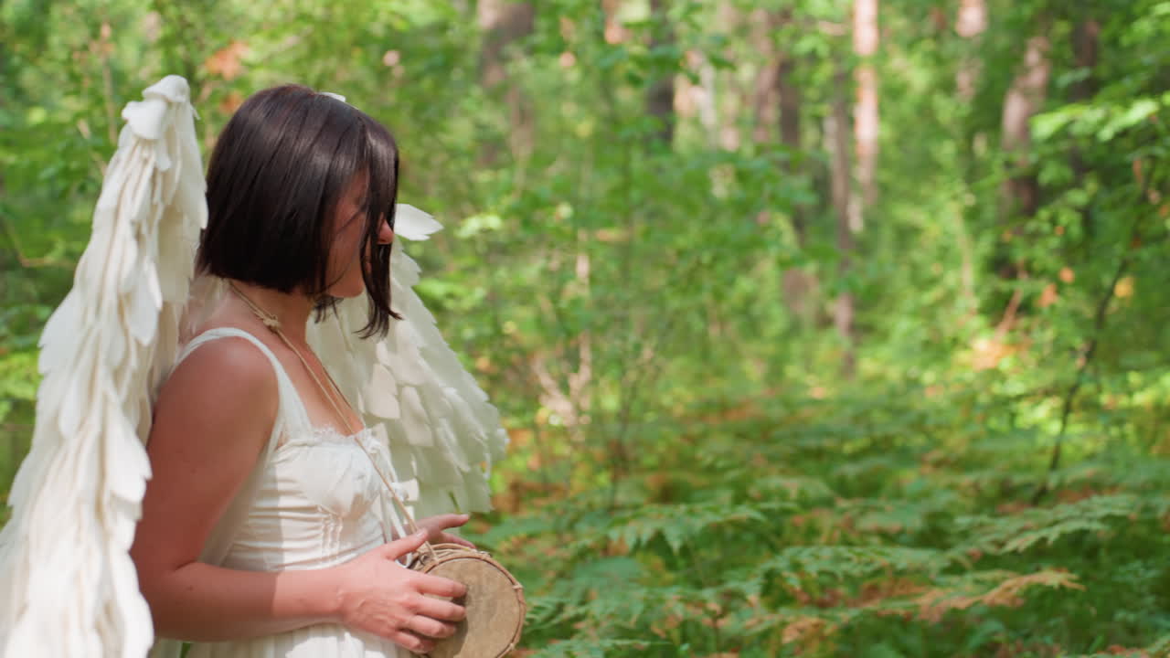 Close up of girl with white angel wings walking through green forest while gently beating small drum, sunlight filtering through leaves, peaceful rhythm echoing nature