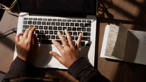 high angle shot of an asian woman typing notes on her laptop computer keyboard next to a notebook in the afternoon light at her dining room table