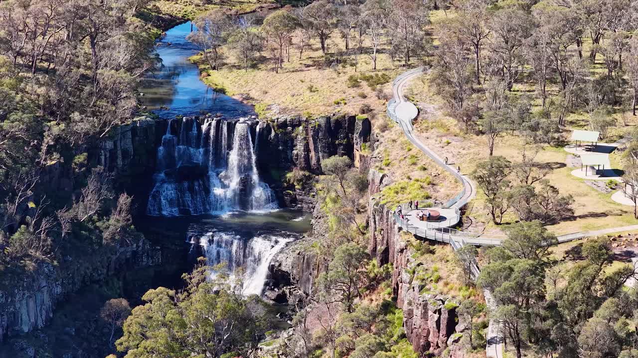 Drone footage glides above a scenic clifftop lookout, revealing a dramatic multi-tiered waterfall, winding walkway, and lush Australian bushland in bright daylight