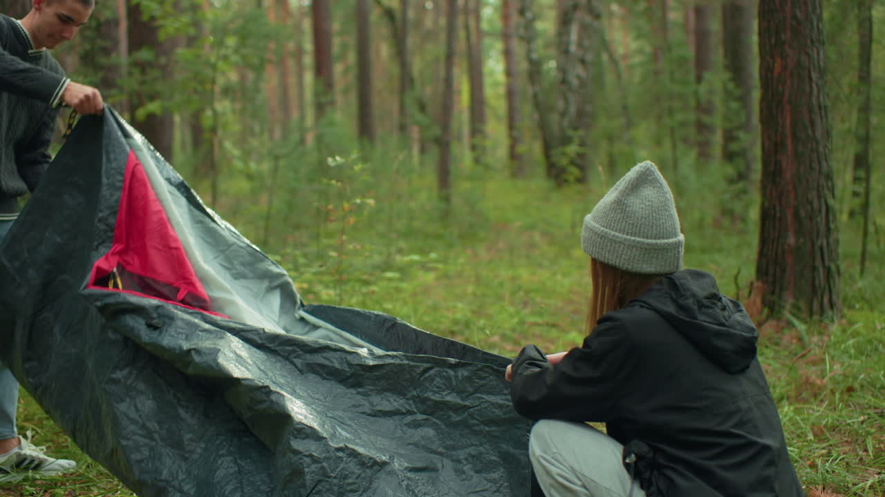 Couple collaborates to pack tent in peaceful forest setting, unfolding fabric while kneeling on ground, sharing affectionate moment as they draw close surrounded by trees