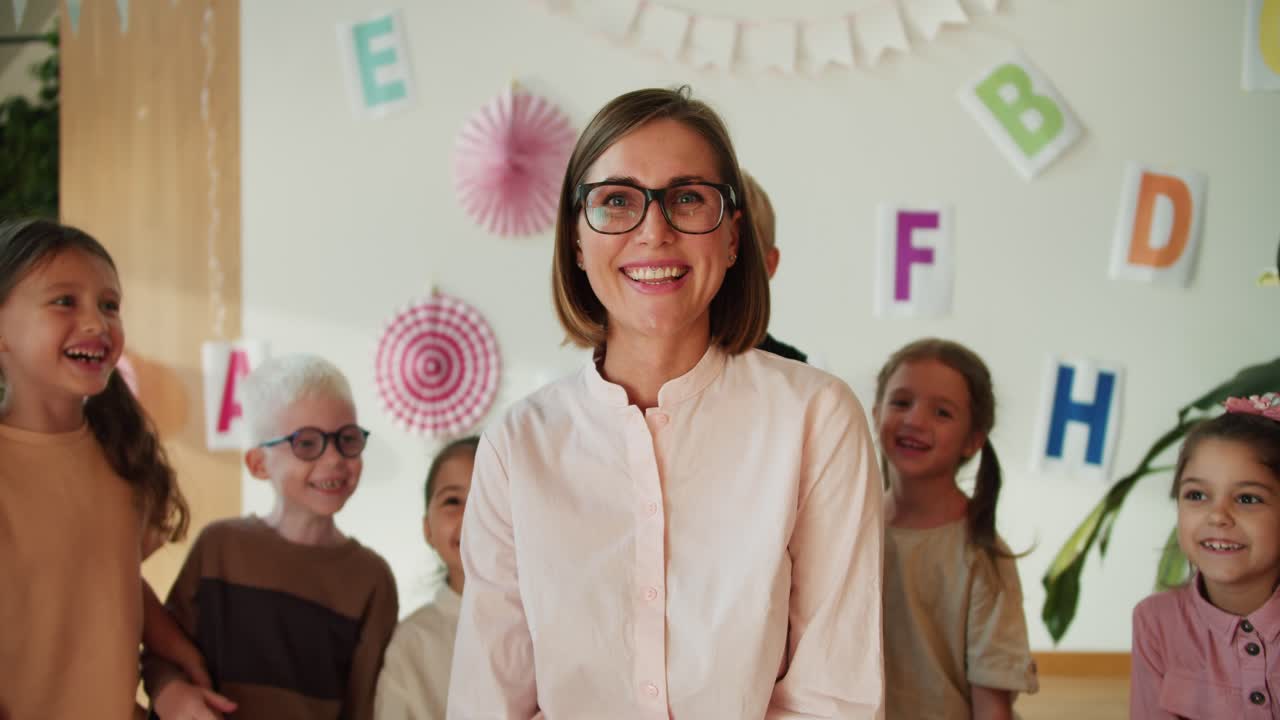 retrato de una feliz maestra con un peinado de bob en gafas y una camisa blanca que mira a la cámara y sonríe ampliamente contra el fondo de un grupo de niños en un club de preparación escolar
