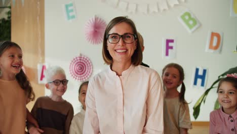 Retrato-De-Una-Profesora-Feliz-Con-Un-Peinado-Bob,-Gafas-Y-Una-Camisa-Blanca-Que-Mira-A-La-Cámara-Y-Sonríe-Ampliamente-En-El-Contexto-De-Un-Grupo-De-Niños-En-Un-Club-De-Preparación-Escolar.