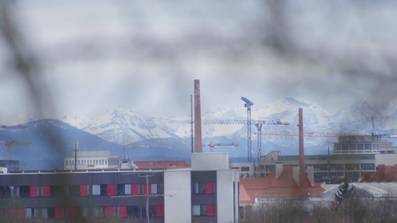 A stunning view of Munich’s urban landscape with construction cranes and industrial buildings in the foreground, contrasted by the snow-capped Alps in the background.