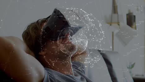 globe of network of connections over caucasian man wearing vr headset sitting on the couch at home