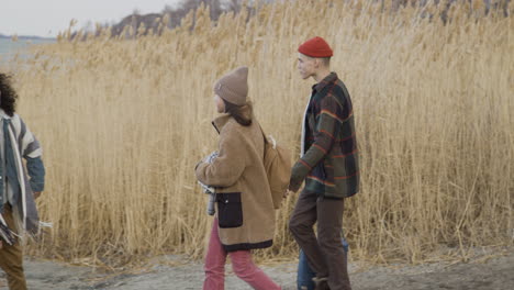 seitenansicht eines jungen und mädchens im teenageralter, die sich zwei freunden in winterkleidung nähern, die an einem bewölkten tag in einem weizenfeld in der nähe des meeres spazieren gehen