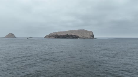 Aerial-Dolly-Drohne-Weitschuss-über-Dem-Meer-Auf-Einer-Insel-Mit-Felsen-Und-Gebäuden-Darauf-An-Der-Küste-Oder-Am-Ufer-Bei-Ruhigen-Wellen-Mit-Booten-Im-Wasser