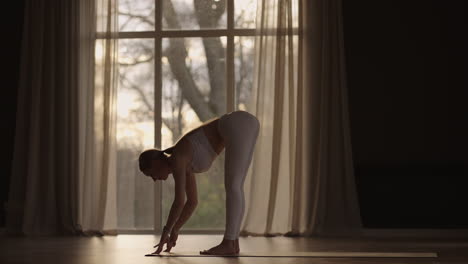 slow motion: young woman is doing yoga in a white room filled with light the girl performs yoga stands and elements near the large window.