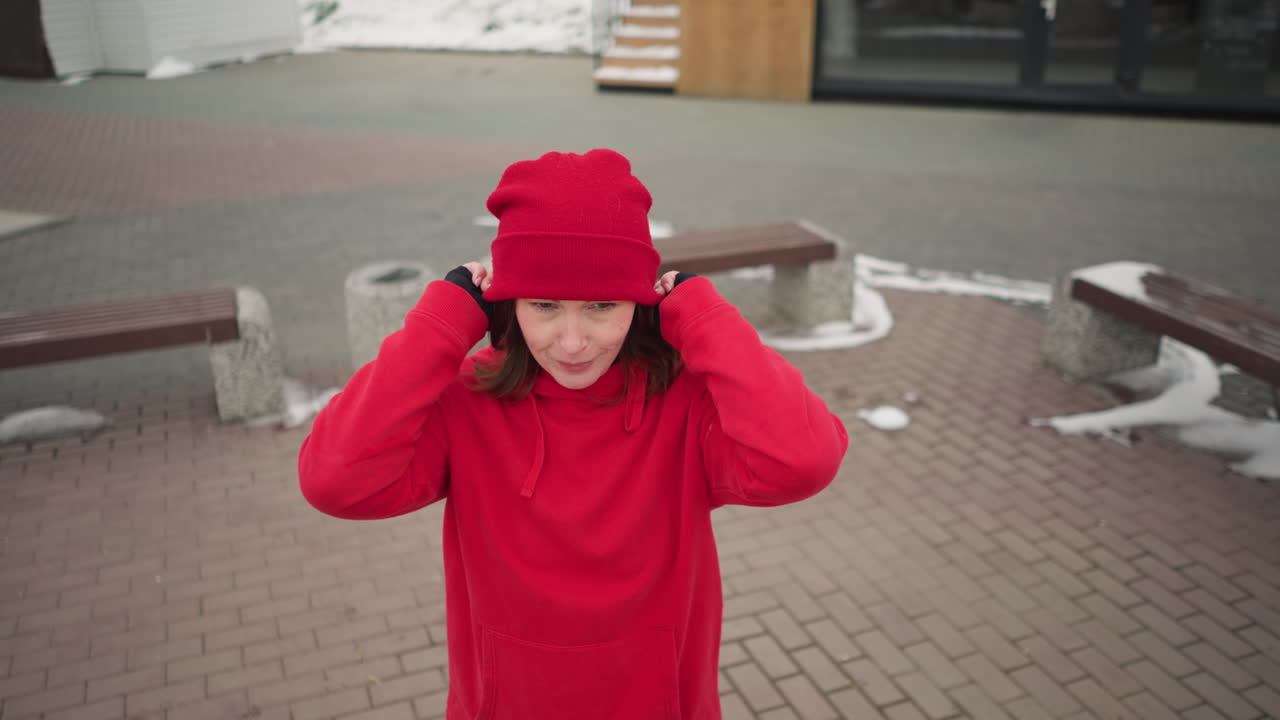 freelancer se pone un gorro rojo al aire libre con una sonrisa cálida durante el invierno, el fondo presenta paisajes urbanos cubiertos de nieve, estructuras de madera, bancos modernos y una atmósfera serena de invierno