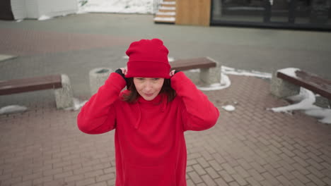 freelancer puts on red beanie outdoors with a warm smile during winter, background features snow-covered urban landscape, wooden structures, modern benches, and a serene winter atmosphere