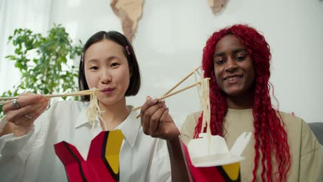 diverse female friends eating noodles together at home using chopsticks, positive mood