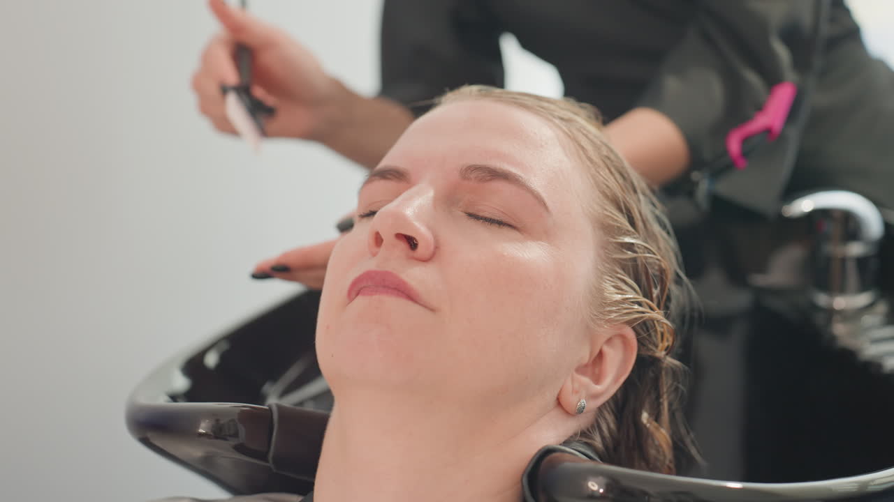 Woman reclining at salon sink receiving gentle scalp treatment from professional stylist using brush and shampoo, hair wet, eyes closed, calm atmosphere, closeup on head and basin, soft lighting