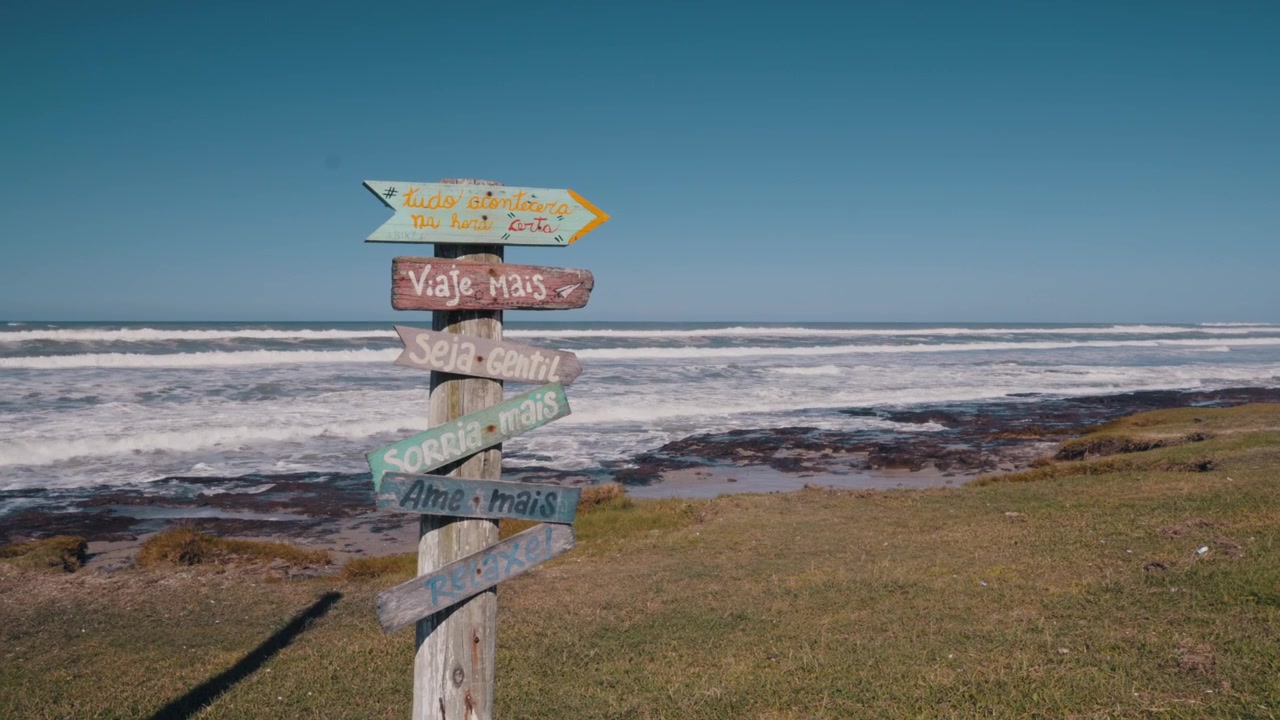 Un cartello di legno con messaggi motivazionali in portoghese si trova su un'area erbosa che si affaccia sull'oceano. Le onde si infrangono dolcemente sullo sfondo sotto un cielo azzurro e limpido, creando un'atmosfera serena e ispiratrice.